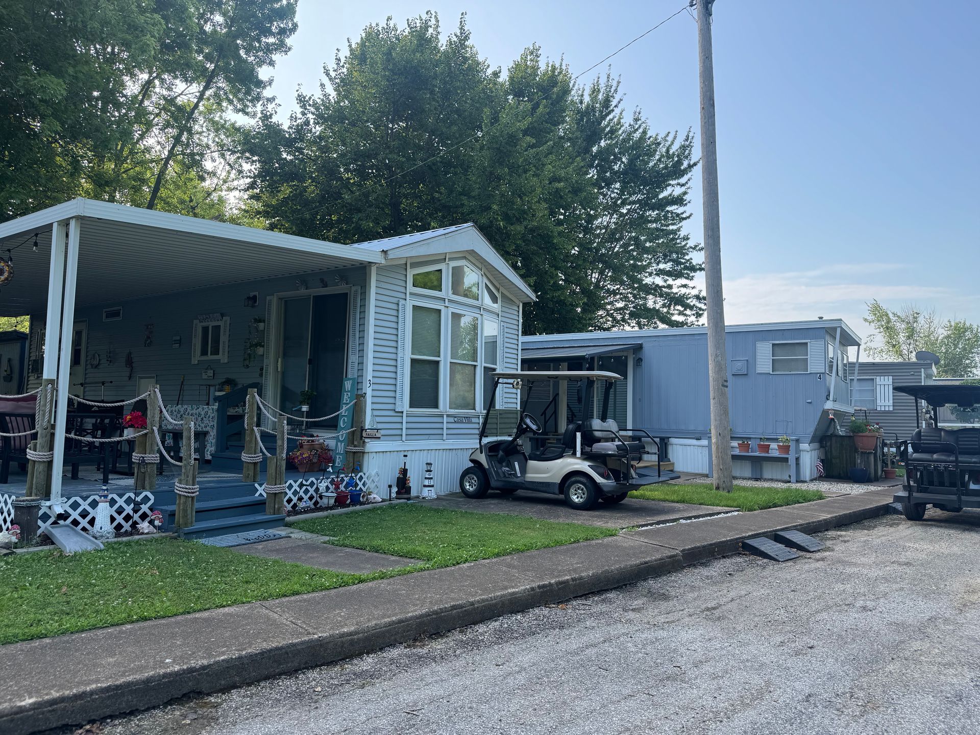 Small white mobile home with porch and golf cart beside a gravel lot under trees and blue sky