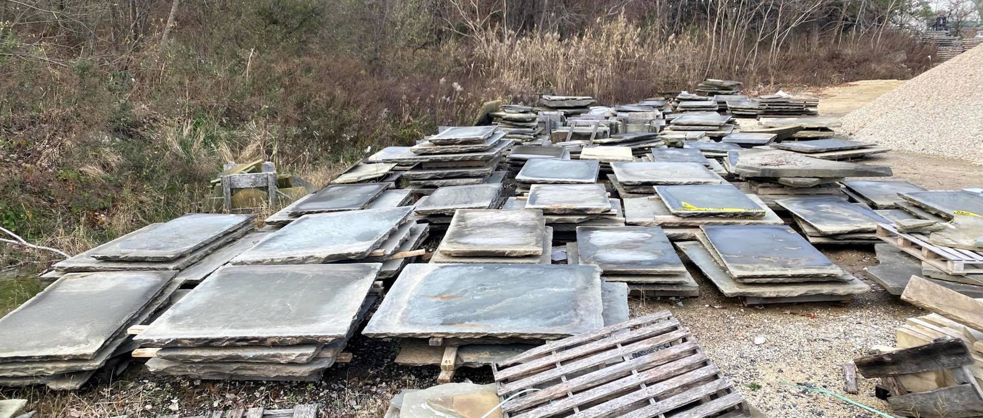 Piles of gray concrete slabs and a wooden pallet are on a gravel surface next to vegetation.