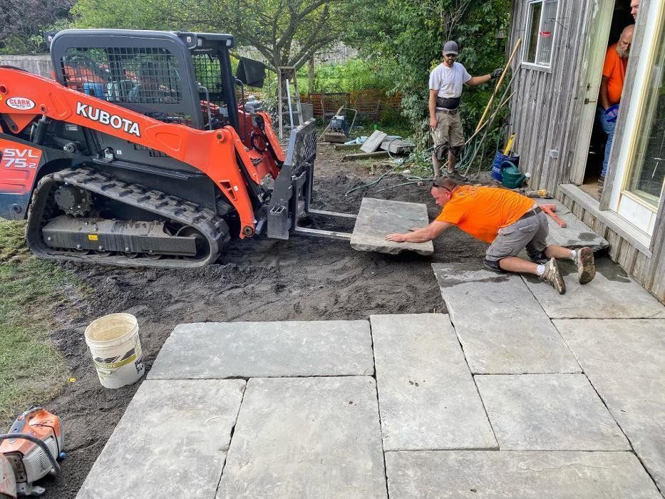 Workers laying stone patio with Kubota skid steer. One person adjusts a slab.