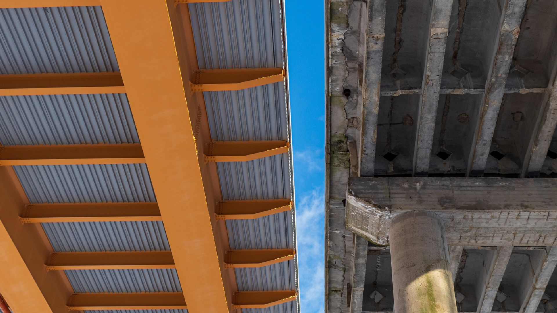 Underside of an orange-and-blue industrial structure beside a gray concrete ceiling with beams and columns
