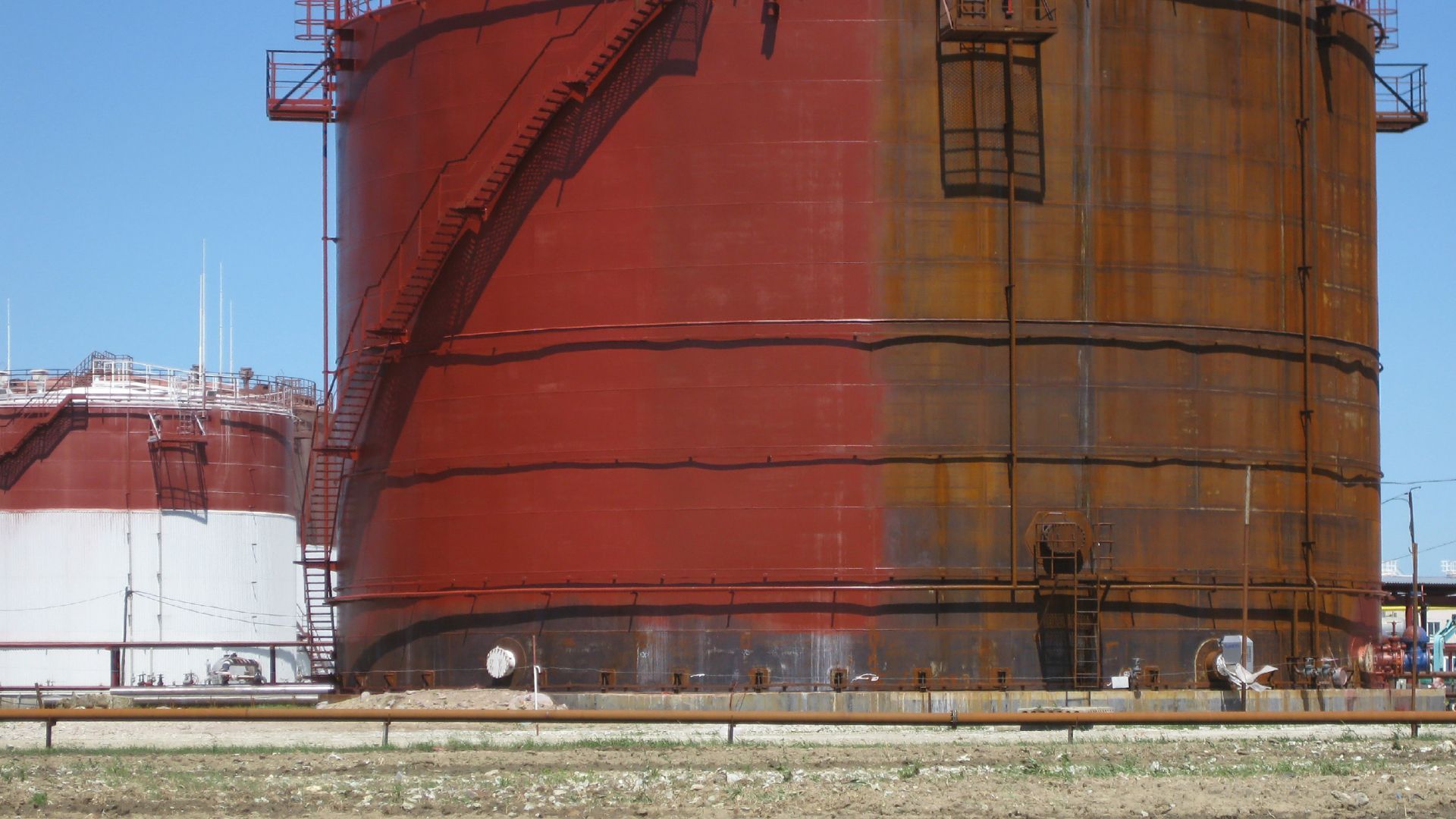 Large rust-red industrial storage tank beside a snowy rail line under a clear blue sky