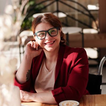 Smiling person in red blazer at a café, leaning on hand beside a coffee cup.