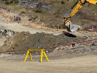 Construction site with excavator digging a dirt embankment behind orange cones and yellow barriers