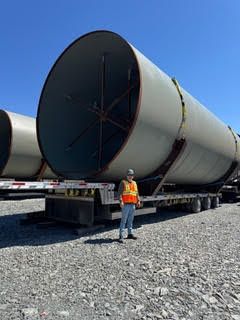 Worker in orange vest beside large cylindrical metal tanks on a flatbed trailer in a gravel yard