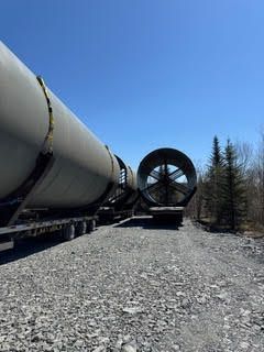 Large industrial pipe sections on a gravel path, with a giant fan-like machinery piece in a clear outdoor yard.