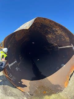 Large rusted metal pipe section on the ground, with a worker partially visible at left under a blue sky.