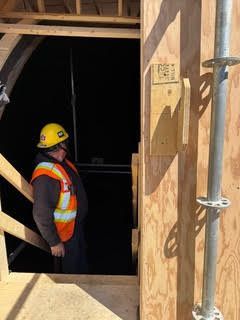 Worker in yellow hard hat and orange vest standing in a dark construction opening with wooden framing.