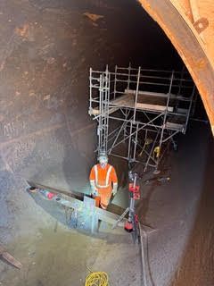 Worker in orange safety vest on scaffolding inside a large tunnel or pipe.