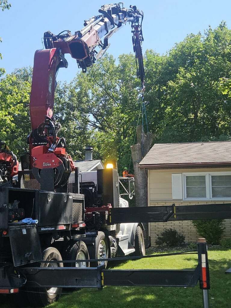 Utility truck with a crane lifting a long black pole beside a suburban house and trees