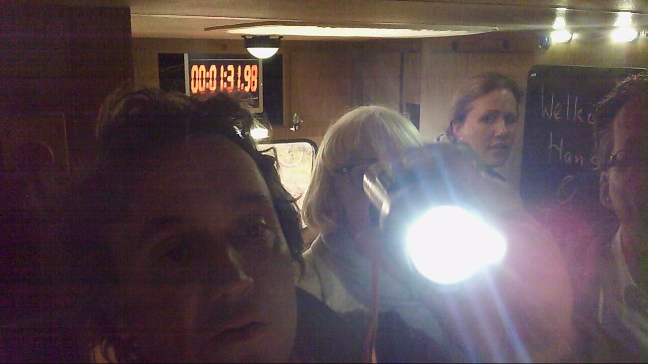 Mensen bevinden zich in een schemerig verlichte ruimte, waar een fel licht op schijnt. Een timerdisplay is zichtbaar.