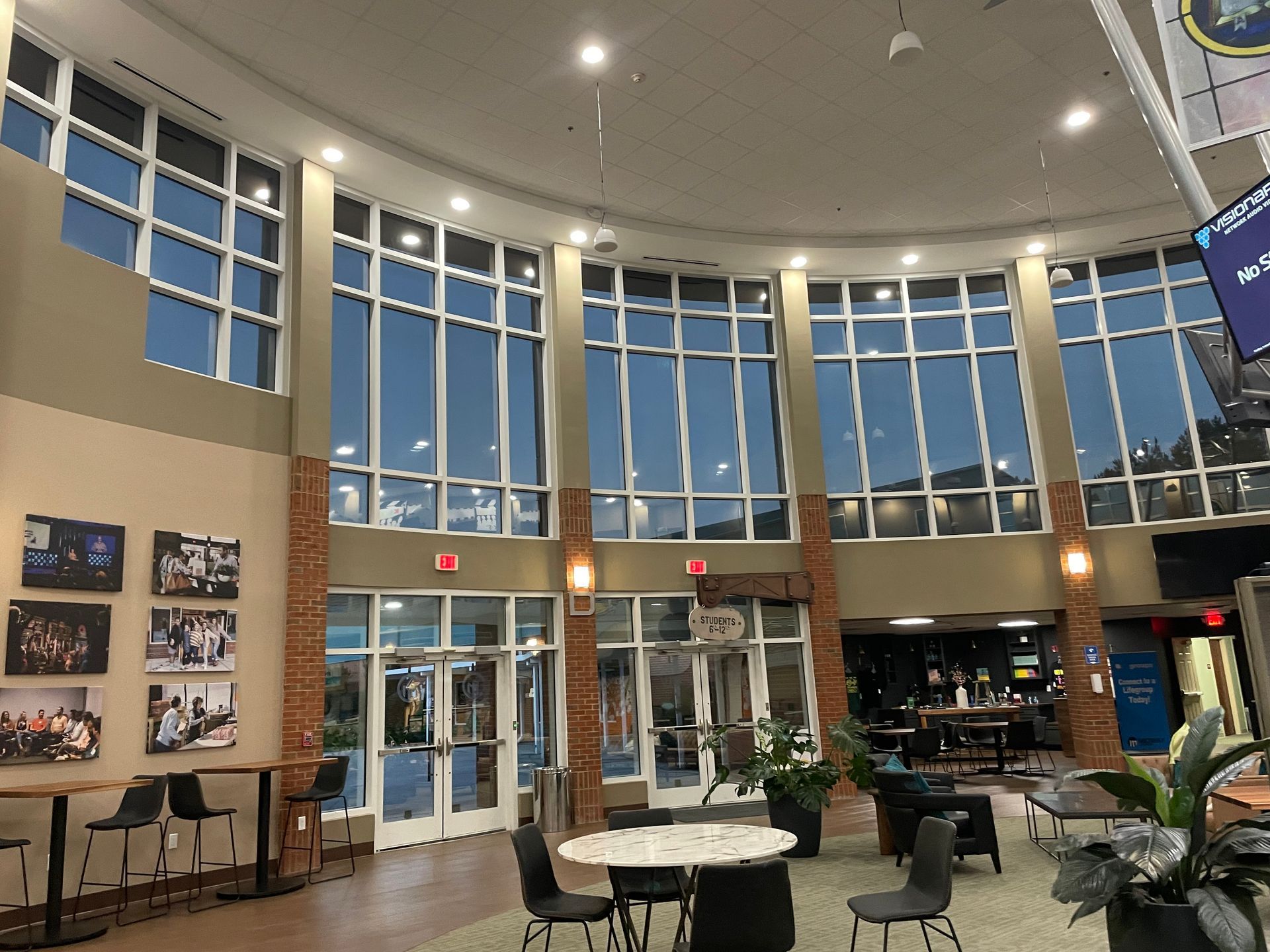 Modern lobby with tall windows, glass entrance, tables, chairs, and wall photo displays at night