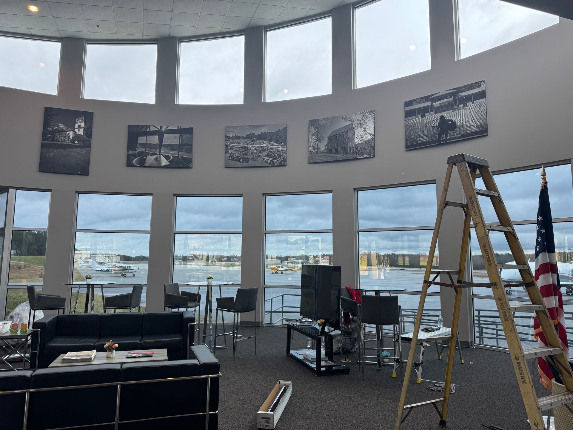 Interior of a waterfront venue with large windows, a ladder, and an American flag; setup in progress.