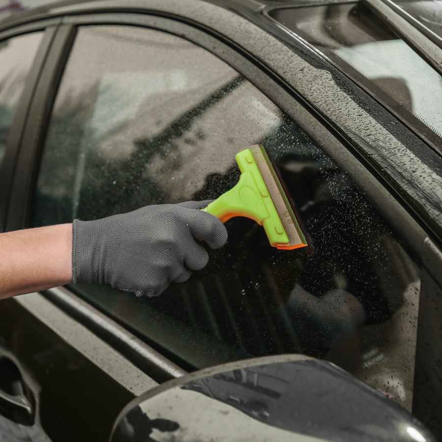 Gloved hand using a green squeegee to clean a car window with water droplets