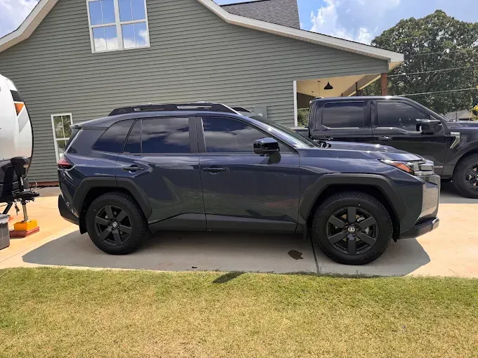 Dark blue SUV parked in a driveway beside a gray house, with another black truck nearby.