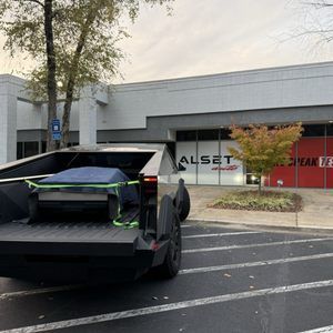 Black Tesla Cybertruck parked outside a modern building with storefront windows and a tree in the lot