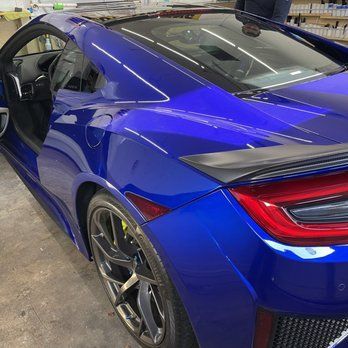 Blue sports car shown from the rear-left with the driver’s door open in a garage.
