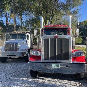 Two parked semi trucks, a red one in front and a white one behind, on a gravel lot under trees
