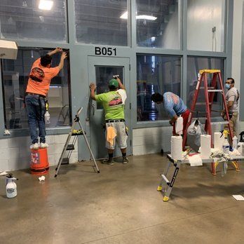 Workers on ladders and buckets cleaning a glass storefront labeled B051 in a warehouse-like space