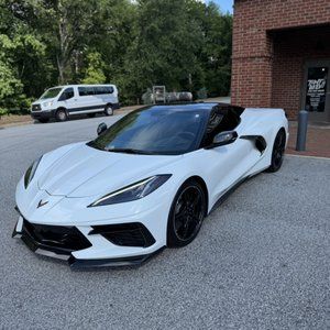 White Chevrolet Corvette parked beside a brick building, with a white van and trees in the background