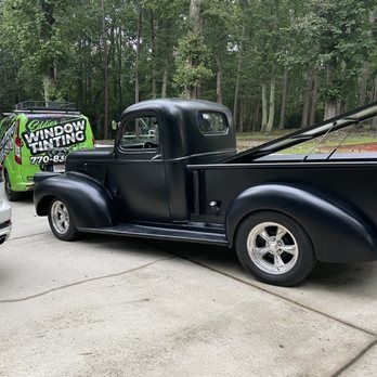 Black vintage pickup truck parked on a driveway beside a green window tinting van in a wooded area