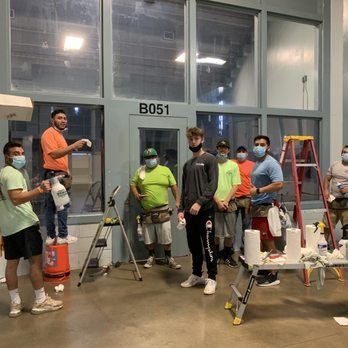 Workers in safety gear posing in a workshop, with tools, ladders, and the label B051 on the wall.