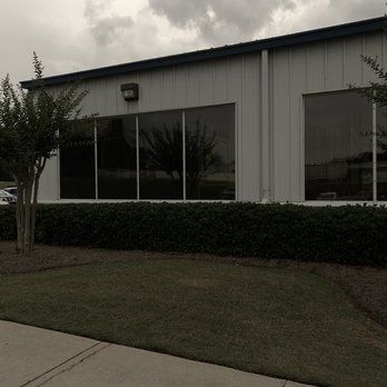 Low industrial building with large windows, trimmed hedges, and a cloudy sky.