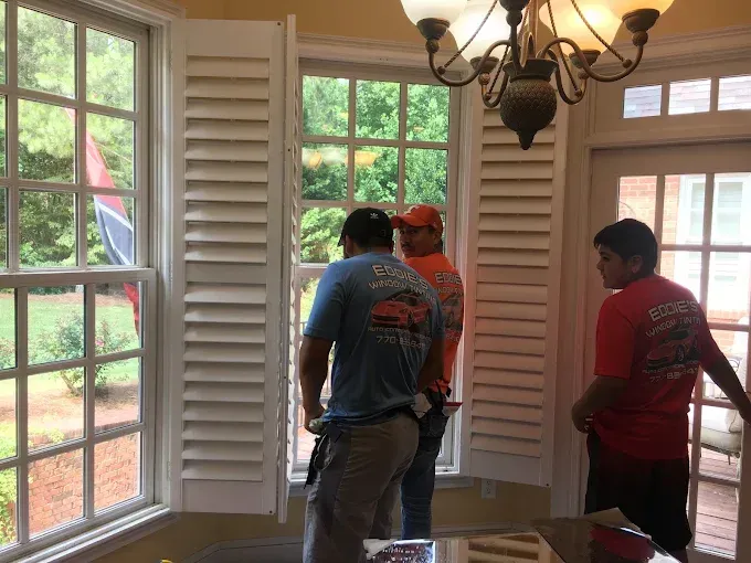 Three workers installing a window treatment in a bright room with large windows and white shutters