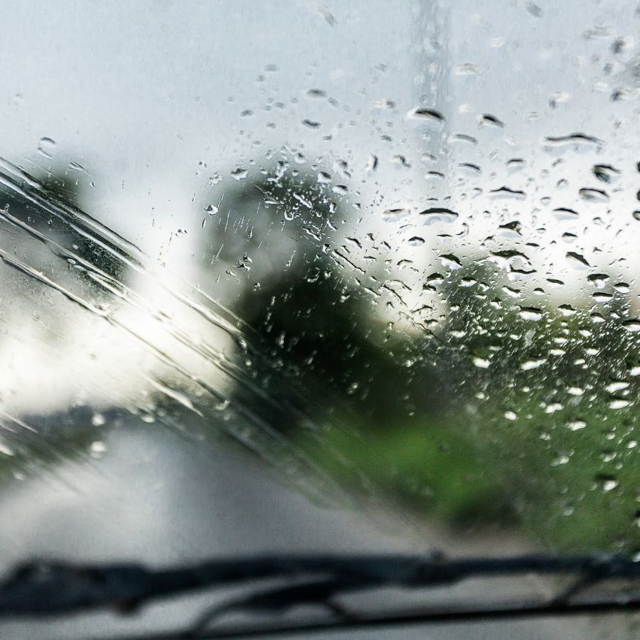 Rain-covered windshield with blurred road and trees beyond the wipers