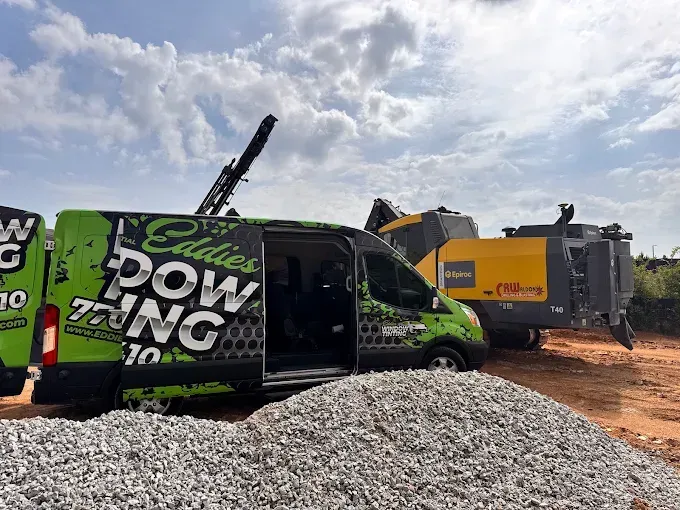 Green utility van beside large yellow construction machine on a dirt site under a cloudy sky.
