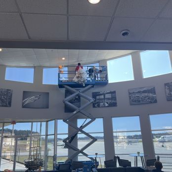 People on a scissor lift inside a bright waterfront room with large windows and wall photos
