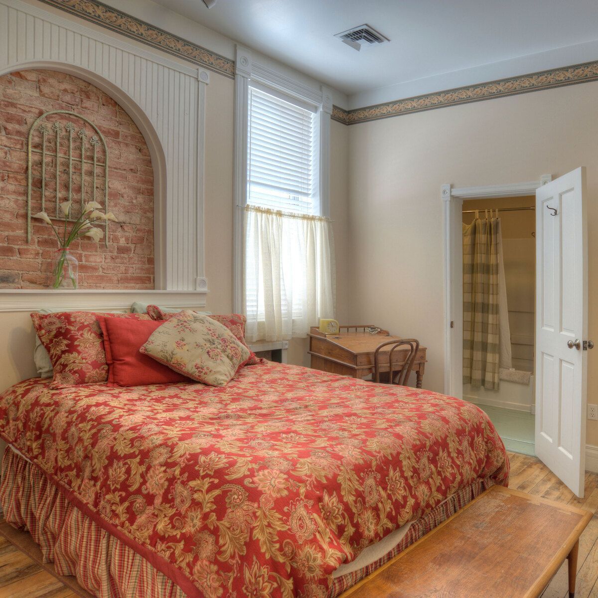 A wide view of a guest room with a red and gold bedspread, wooden furniture, and a brick wall decoration.