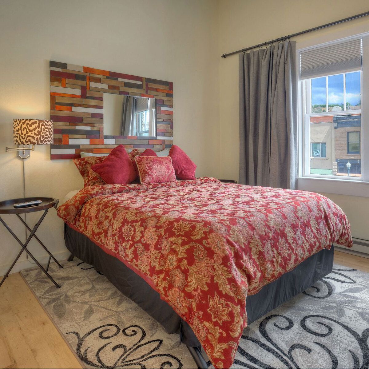 A neatly made bed with a red and gold floral patterned comforter and pillows, with a window and artwork on the wall.