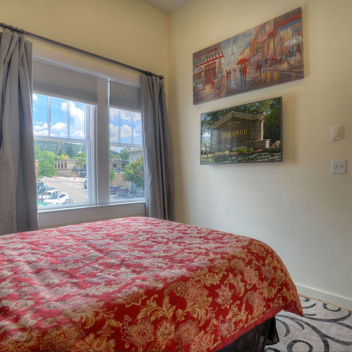 A room with a red and gold bedspread, cream walls, and two pieces of artwork.