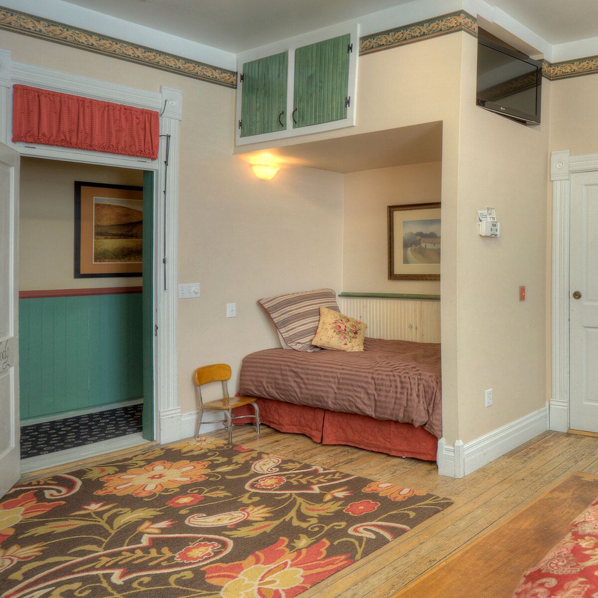 A small hotel room with a single bed, floral rug, and TV on the wall.