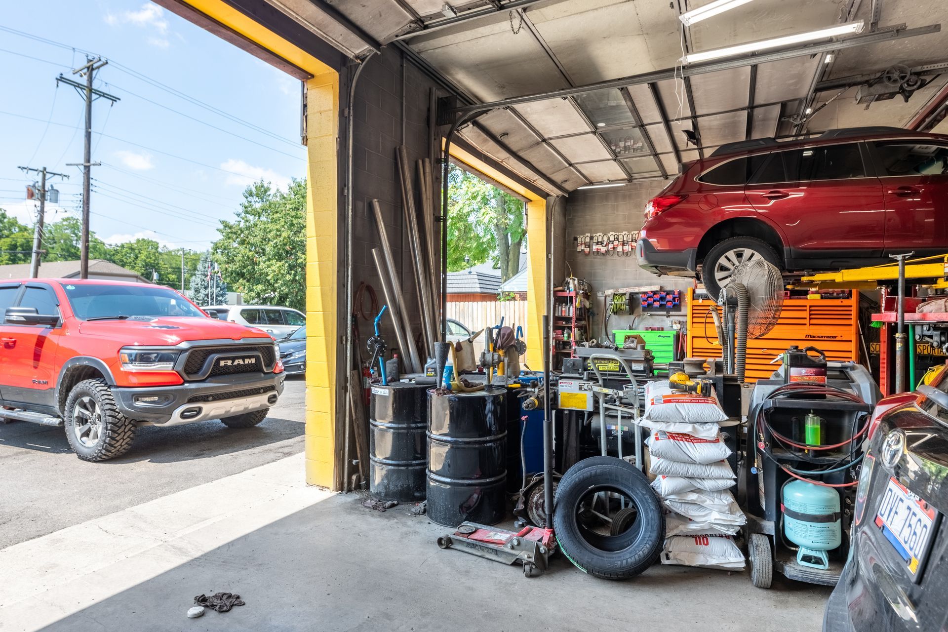 An auto repair shop: a red truck parked outside, a car on a lift inside, tools, barrels, and open garage door.