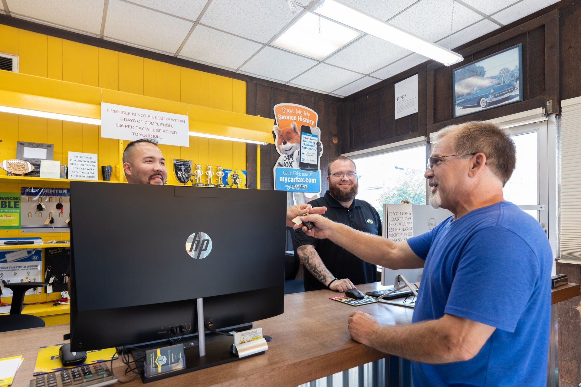 Man at counter with two employees in a shop; one points at a computer monitor. Yellow wall in the background.