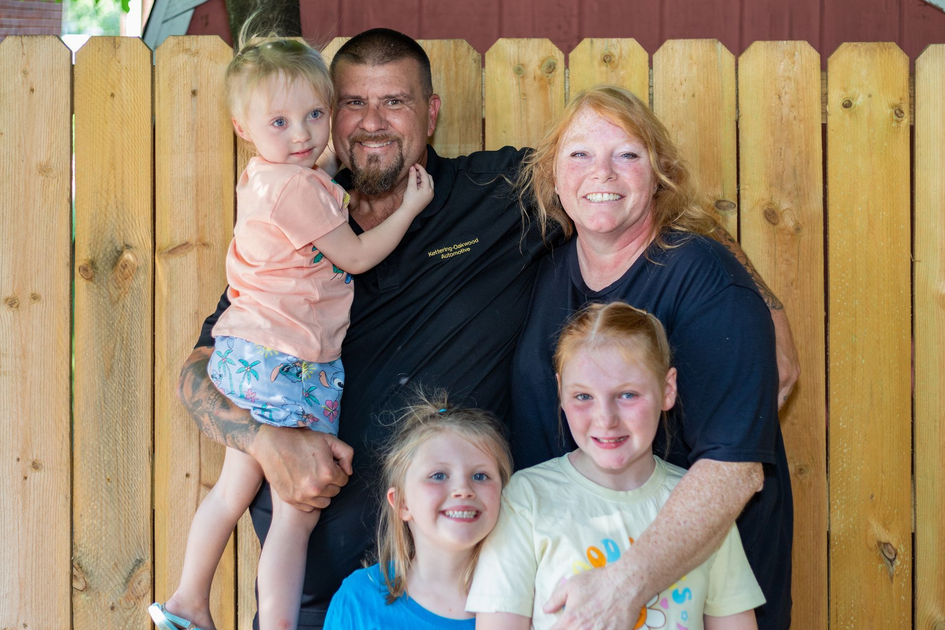 Family of five smiling, posing in front of a wooden fence. Man holds a child, woman hugs two children.