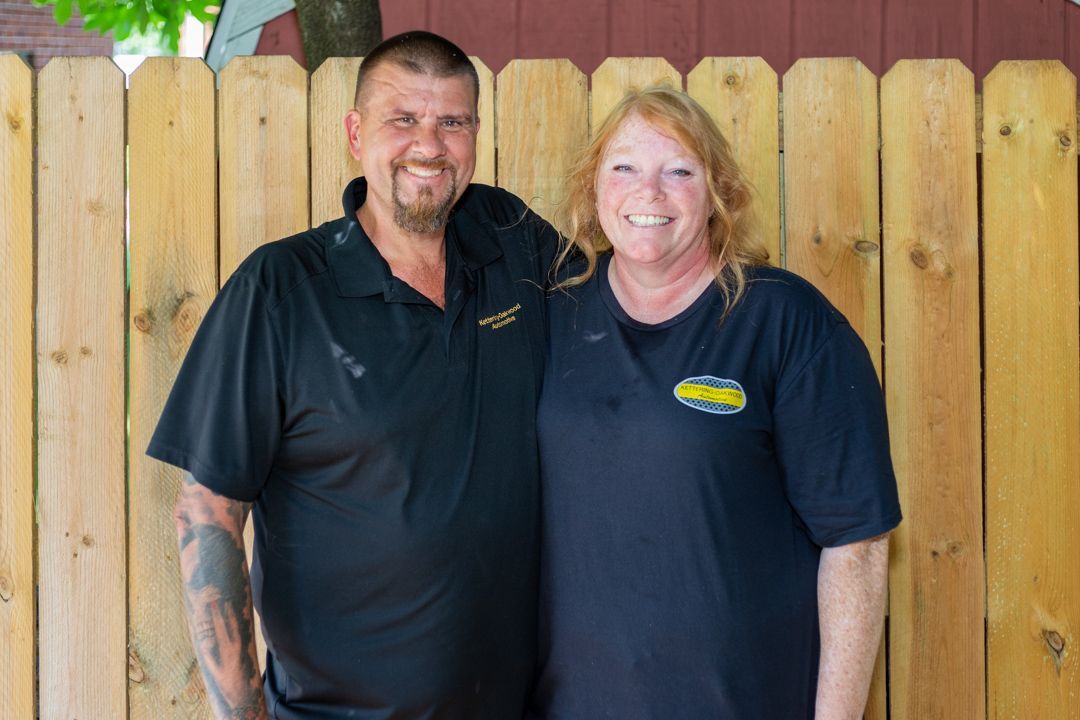 Two people smiling in front of a wooden fence. Man in black polo has a tattoo, woman in black tee has red hair.