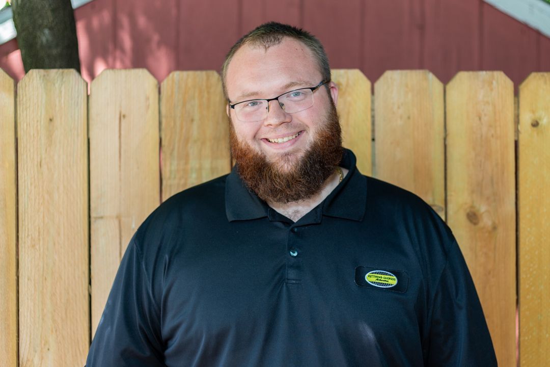 Man with glasses and a red beard, wearing a black shirt, smiles in front of a wooden fence.