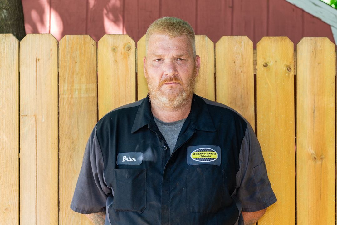 Man in a mechanic uniform stands in front of a wooden fence.