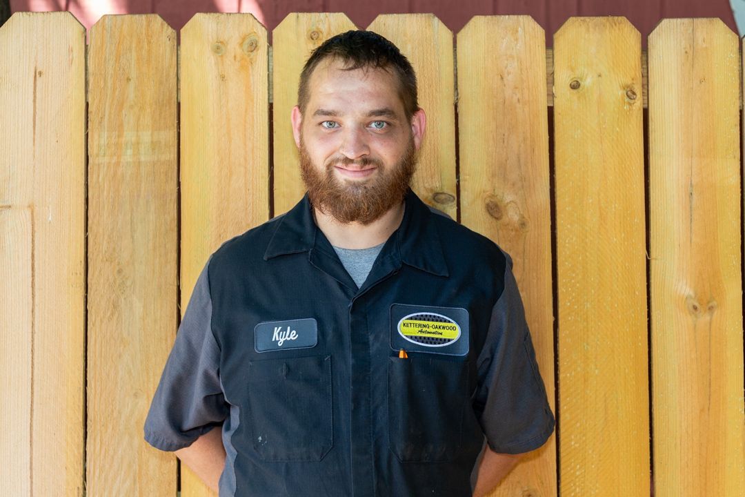 Man with brown beard in mechanic uniform smiles in front of a wood fence.