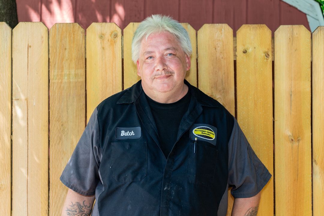 Man with white hair in work uniform stands in front of a wooden fence.