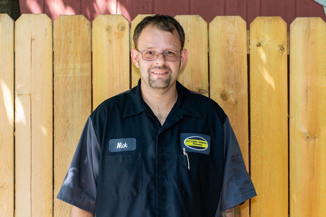 Man in mechanic's uniform stands in front of a wooden fence, smiling.