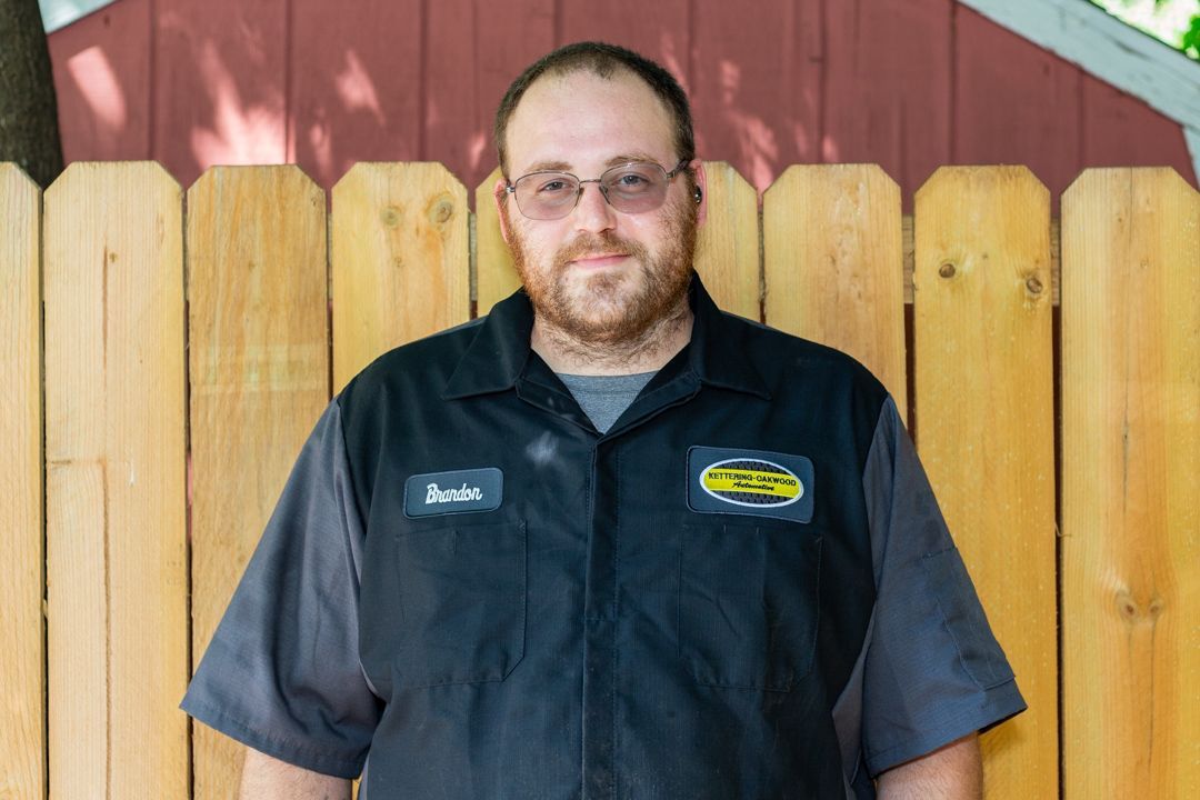 Man wearing glasses and mechanic's shirt stands in front of a wooden fence.