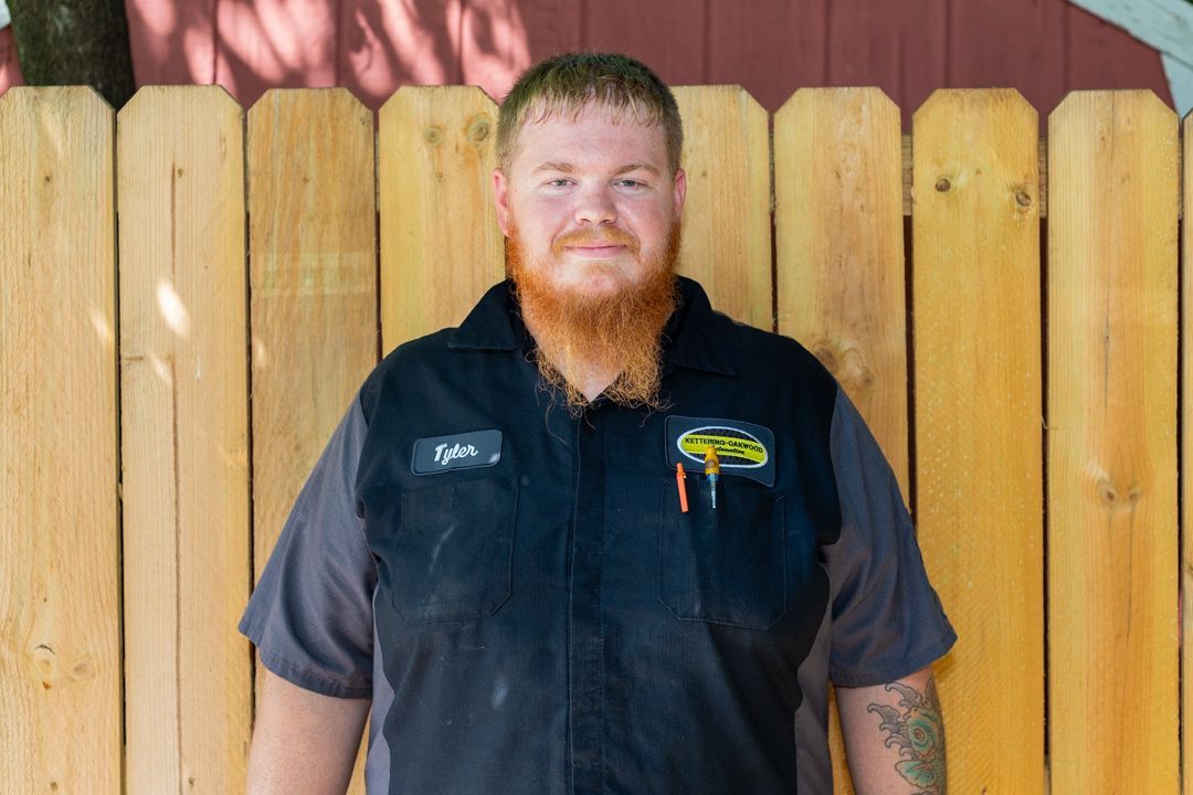 Man with red beard, wearing a mechanic uniform, stands in front of a wooden fence.