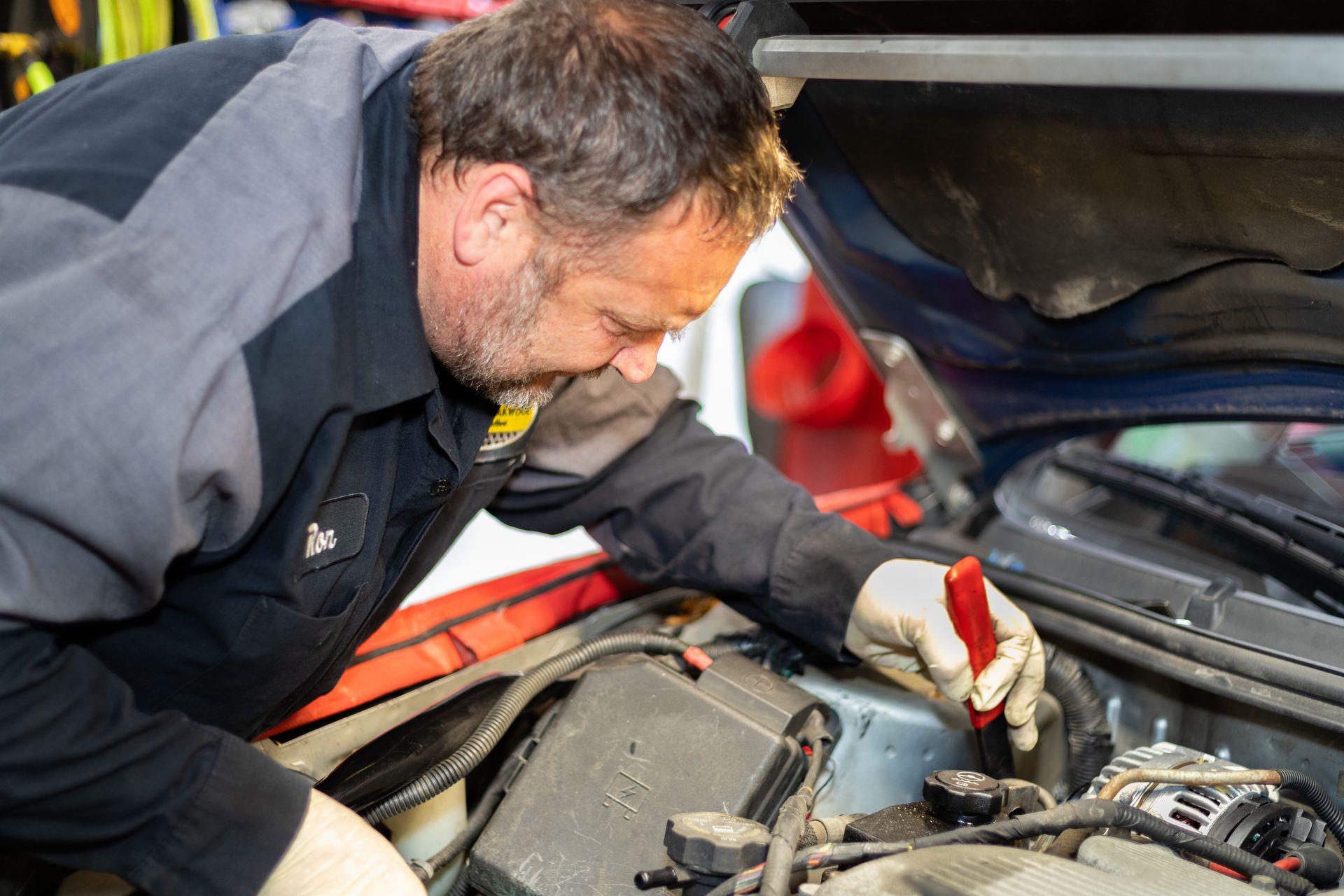 Mechanic working on a car engine under the hood, using a red tool.