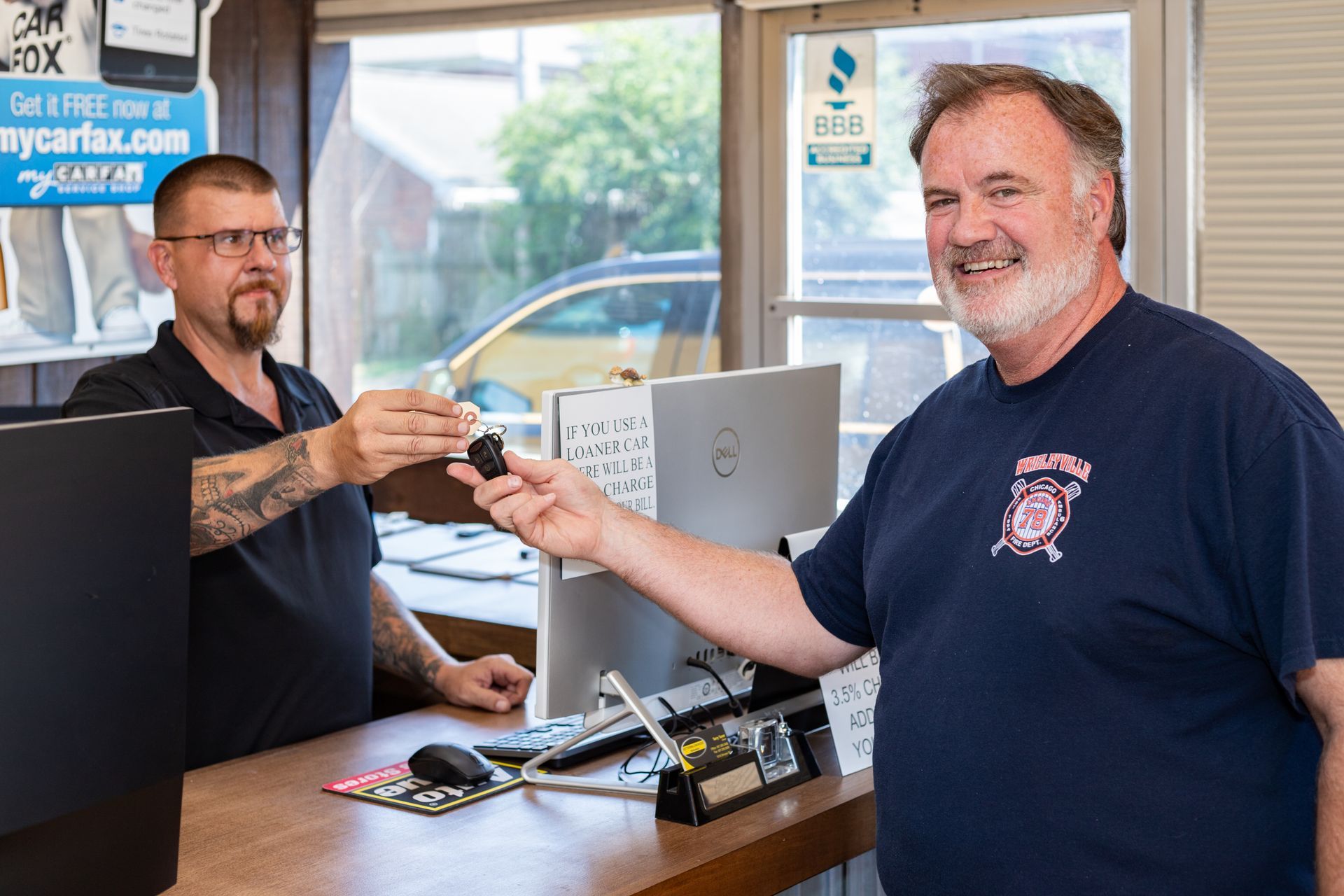 Two men at a car repair shop. One hands keys to the other. Counter, computer, and car outside visible.