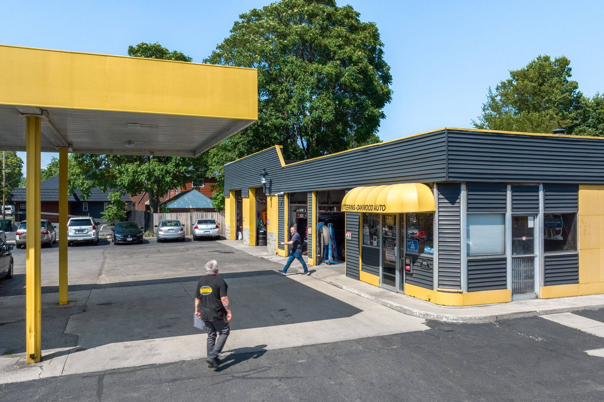 Yellow-trimmed building with a canopy. People walk around the shop and parking area on a sunny day.