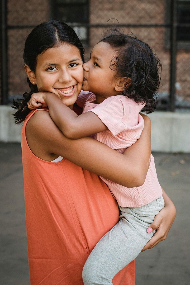 Two children hugging outdoors, one smiling while carrying the other in pink shirt and gray leggings.