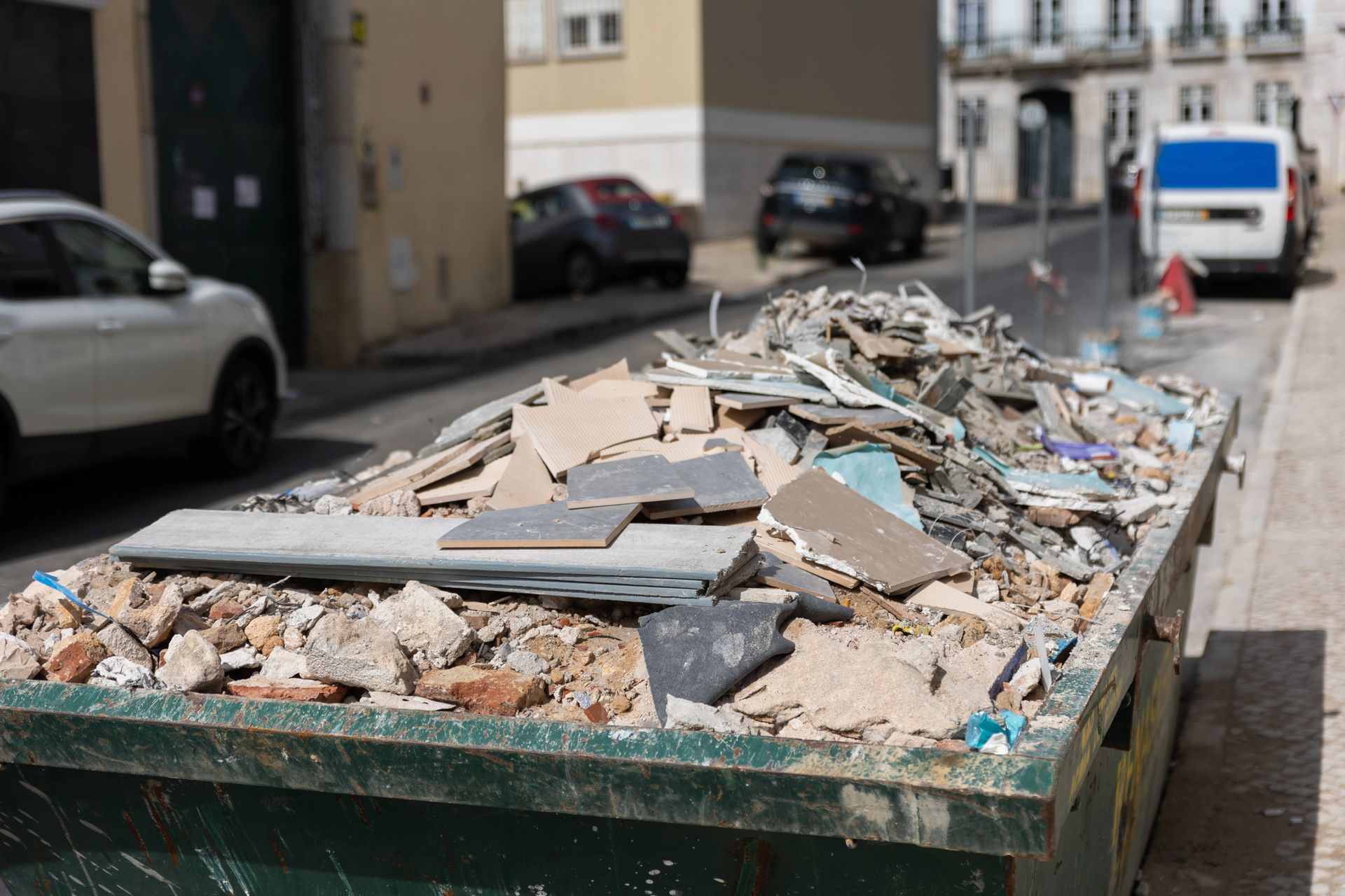 Detriti edili ammucchiati in un cassonetto verde su una strada cittadina, con auto parcheggiate nelle vicinanze.